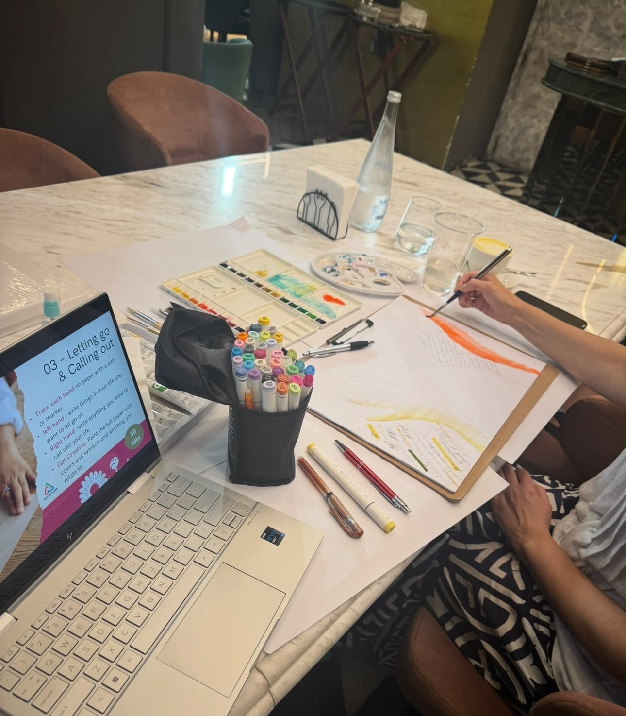 A worked sheet with two hands traced and painted in orange and yellow, watercolour palettes, brushes and marker pens spread across the table beside a laptop